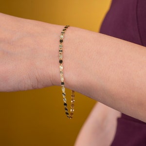 K&ouml;nnte beinhalten: Ein zartes goldenes Armband mit abwechselnd sternf&ouml;rmigen und ovalen Gliedern. Das Armband wird am Handgelenk einer Person vor einem gelben und burgunderfarbenen Hintergrund pr&auml;sentiert. Der Schmuck ist ein schlichtes, aber elegantes Accessoire.