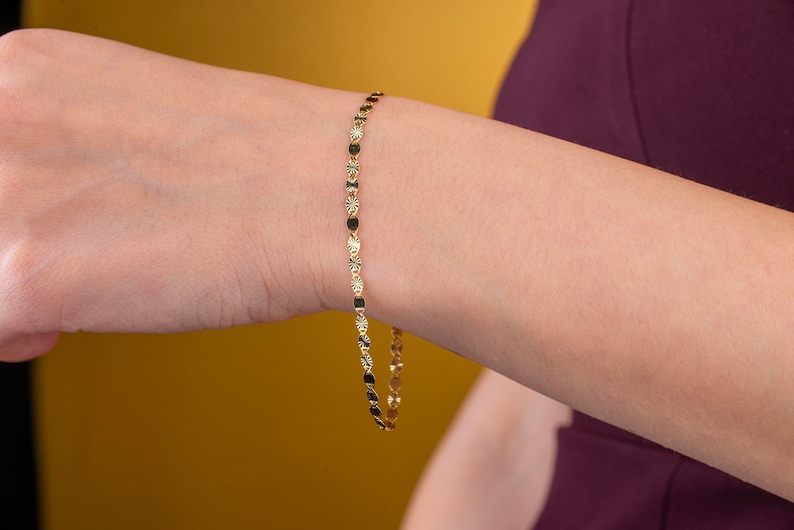 K&ouml;nnte beinhalten: Ein zartes goldenes Armband mit abwechselnd sternf&ouml;rmigen und ovalen Gliedern. Das Armband wird am Handgelenk einer Person vor einem gelben und burgunderfarbenen Hintergrund pr&auml;sentiert. Der Schmuck ist ein schlichtes, aber elegantes Accessoire.