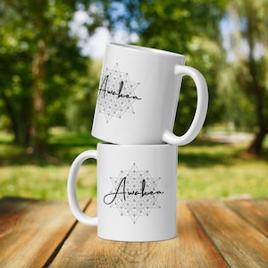Könnte beinhalten: Zwei weiße Keramikbecher, die übereinander gestapelt sind. Beide Becher haben ein geometrisches Design mit dem Wort "Awaken" in schwarzer Kursivschrift.
