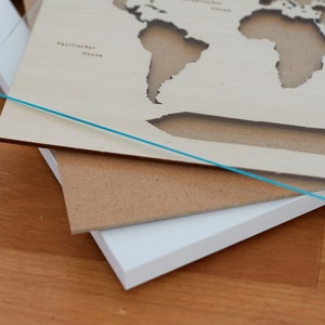 May include: A wooden world map with cut-out continents, layered on top of a brown and white mat. The map is made of light wood and has the words "Pazifischer Ozean" and "Indischer Ozean" printed on it.