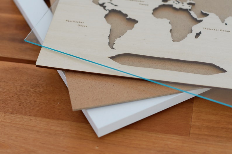 May include: A wooden world map with cut-out continents, layered on top of a brown and white mat. The map is made of light wood and has the words "Pazifischer Ozean" and "Indischer Ozean" printed on it.