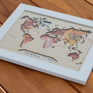 May include: A white framed wooden world map with cutouts for each continent. The cutouts are filled with various currency bills. The map is signed "Marc & Miriam 2.2.2022".