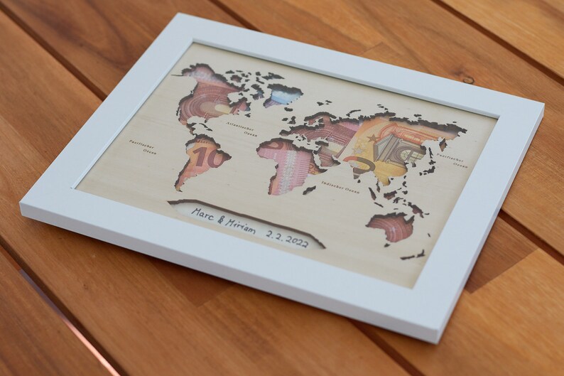 May include: A white framed wooden world map with cutouts for each continent. The cutouts are filled with various currency bills. The map is signed "Marc & Miriam 2.2.2022".