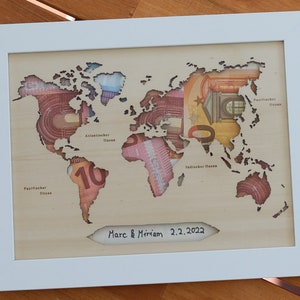 May include: A wooden world map with a cutout design, featuring euro banknotes inside. The map is framed in white and has the inscription "Marc & Miriam 2.2.2022" at the bottom.
