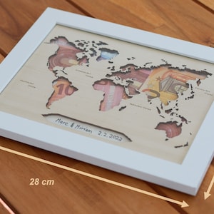 May include: A white framed wooden world map with cutouts for each continent. The cutouts are filled with currency from different countries. The map is signed "Marc & Miriam 2.2.2022" and measures 28 inches by 22 inches.