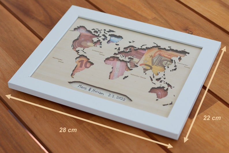May include: A white framed wooden world map with cutouts for each continent. The cutouts are filled with currency from different countries. The map is signed "Marc & Miriam 2.2.2022" and measures 28 inches by 22 inches.