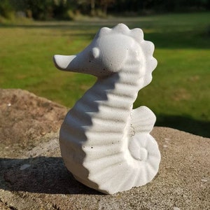 May include: A concrete seahorse sculpture in a light gray color. The seahorse is detailed with textured ridges and a curled tail. The sculpture is placed on a stone surface with a green grassy background.