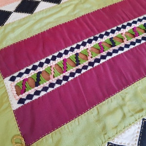 May include: A close-up of a multi-coloured fabric with a geometric pattern. The fabric is made of different shades of pink, green, black, and white. The fabric is sewn together with a decorative stitch.