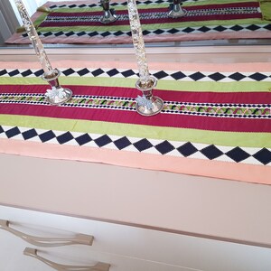 May include: A colourful table runner with a black and white diamond pattern, red and green stripes, and gold tassels. The runner is on a light brown surface with two silver candle holders.