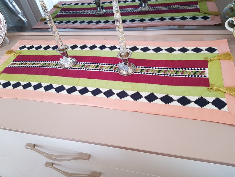 May include: A colourful table runner with a black and white diamond pattern, red and green stripes, and gold tassels. The runner is on a light brown surface with two silver candle holders.