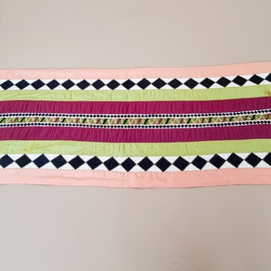 May include: A pink, green, burgundy, black and white table runner with a diamond pattern and tassels on the ends.