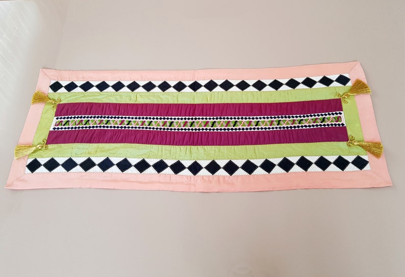 May include: A pink, green, burgundy, black and white table runner with a diamond pattern and tassels on the ends.