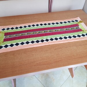 May include: A colourful table runner with a black and white diamond pattern, green and red stripes, and gold tassels on the ends.