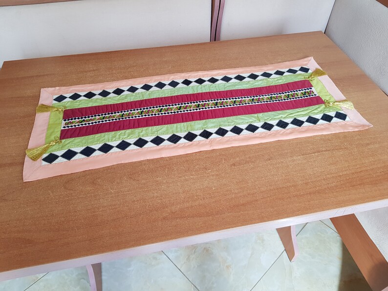 May include: A colourful table runner with a black and white diamond pattern, green and red stripes, and gold tassels on the ends.