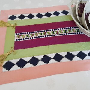 May include: A pink, green, burgundy, and black table runner with diamond patterns and tassels. The runner is made of fabric and has a decorative embroidered design.
