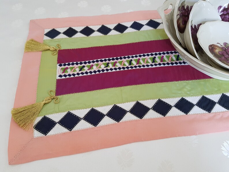 May include: A pink, green, burgundy and black table runner with diamond patterns and tassels. The runner is made of fabric and has a decorative embroidered design.