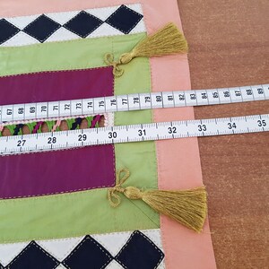 May include: A close-up of a fabric with a patchwork design. The fabric is made up of different coloured squares, including pink, green, and purple. A white measuring tape is laid across the fabric, with the numbers 25 to 36 and 74 to 93 visible.