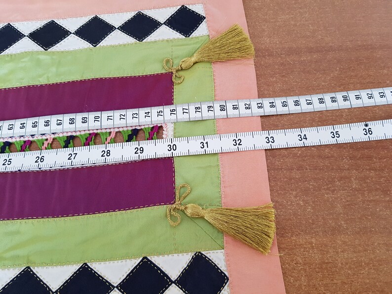 May include: A close-up of a fabric with a patchwork design. The fabric is made up of different coloured squares, including pink, green, and purple. A white measuring tape is laid across the fabric, with the numbers 25 to 36 and 74 to 93 visible.