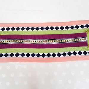 May include: A pink, green and black table runner with a diamond pattern and embroidered details. The runner has tassels on each end.