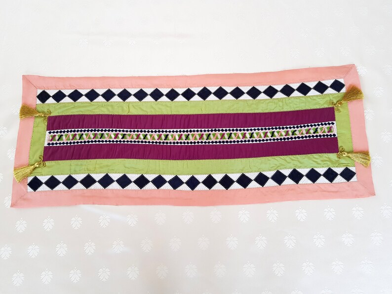 May include: A pink, green and black table runner with a diamond pattern and embroidered details. The runner has tassels on each end.