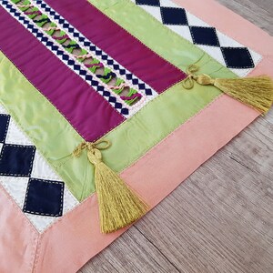 May include: A pink, green, white and black table runner with a diamond pattern and tassels. The runner is made of fabric and has a patchwork design.