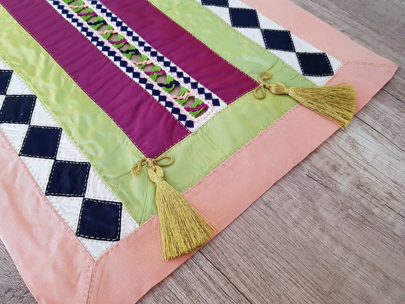May include: A pink, green, white and black table runner with a diamond pattern and tassels. The runner is made of fabric and has a patchwork design.