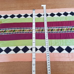 May include: A pink, green, burgundy and black table runner with a diamond pattern and embroidered details. The runner is 94 cm long.