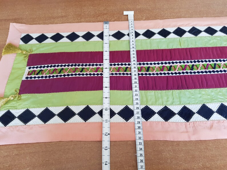 May include: A pink, green, burgundy and black table runner with a diamond pattern and embroidered details. The runner is 94 cm long.
