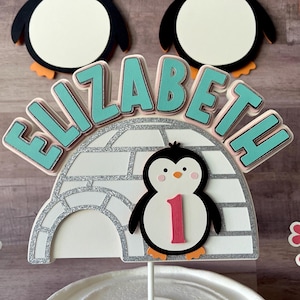 May include: A cake topper featuring a penguin in front of an igloo with the name Elizabeth and the number 1. The topper is decorated with silver glitter and turquoise blue letters.