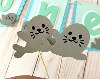 May include: Two gray seal cupcake toppers with black eyes and whiskers. The seals are attached to wooden skewers.