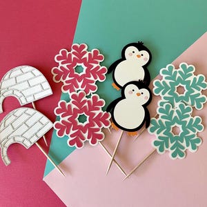 May include: A set of six winter-themed cake toppers. The toppers include two penguins, two snowflakes, and two igloos. The penguins are black and white, the snowflakes are blue and white, and the igloos are white with a silver glitter finish.