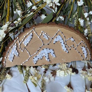 May include: White string art on a wooden slice, depicting a mountain range.