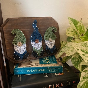 May include: Three gnomes made of string art on a wooden plaque. The gnomes are green, blue, and white. The plaque is sitting on top of a stack of three books. The books are "The Last Song" by Nicholas Sparks, "After the Fire" by Will Hill, and "Broken Things" by  Will Hill.