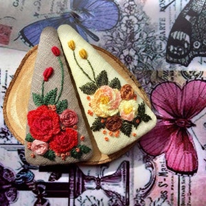 May include: Two embroidered fabric hair clips. One clip is beige with a floral design in shades of pink, yellow, and brown. The other clip is gray with a floral design in shades of red, pink, and green.