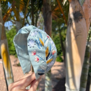 May include: A light blue headband with a floral embroidery design. The embroidery features white, pink, yellow, and blue flowers and leaves.