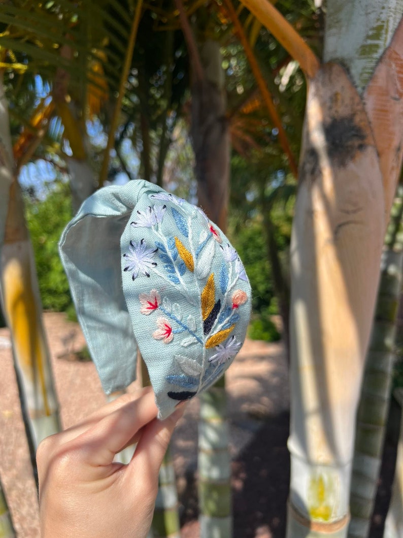 May include: A light blue headband with a floral embroidery design. The embroidery features white, pink, yellow, and blue flowers and leaves.