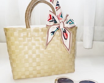 Puede incluir: Un bolso de mano de paja tejido con un asa de color marrón claro y una bufanda estampada en blanco y rosa atada alrededor del asa. Unas gafas de sol con montura transparente y lentes marrones están en primer plano.
