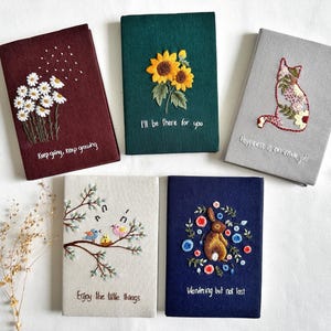 May include: Five small, embroidered notebooks with different designs and inspirational quotes. The notebooks are on a white surface. The quotes are: "Keep going, keep growing", "I'll be there for you", "Happiness is an inside job", "Enjoy the little things", and "Wandering but not lost".
