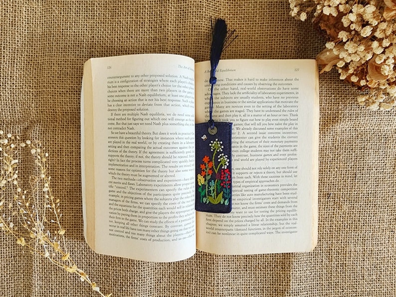 May include: A blue fabric bookmark with colorful embroidered flowers and a white butterfly. The bookmark is folded over a book with text about game theory.
