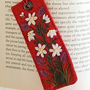 May include: Red fabric bookmark with embroidered white flowers and green leaves. The bookmark has a metal ring at the top.