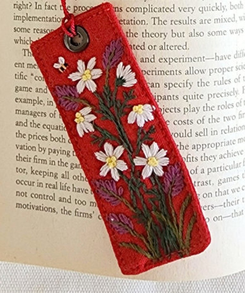 May include: Red fabric bookmark with embroidered white flowers and green leaves. The bookmark has a metal ring at the top.