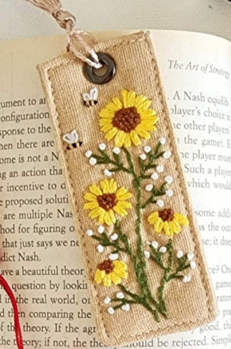May include: A beige fabric bookmark with embroidered yellow sunflowers and white flowers. The bookmark has a brown leather tassel and a metal ring.