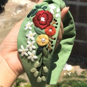 May include: A green fabric headband with a knotted bow and embroidered flowers in red, white, yellow, and orange. The headband is decorated with pearl-like beads.