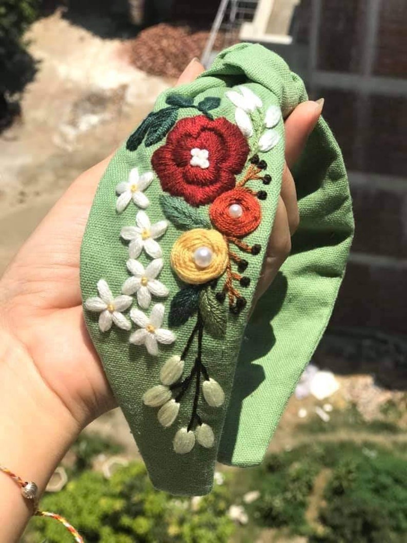 May include: A green fabric headband with a knotted bow and embroidered flowers in red, white, yellow, and orange. The headband is decorated with pearl-like beads.