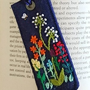 May include: A dark blue fabric bookmark with a metal ring at the top. The bookmark is embroidered with a variety of colorful flowers, including red, orange, yellow, green, and white. A small white bird with a black bee is also embroidered on the bookmark.
