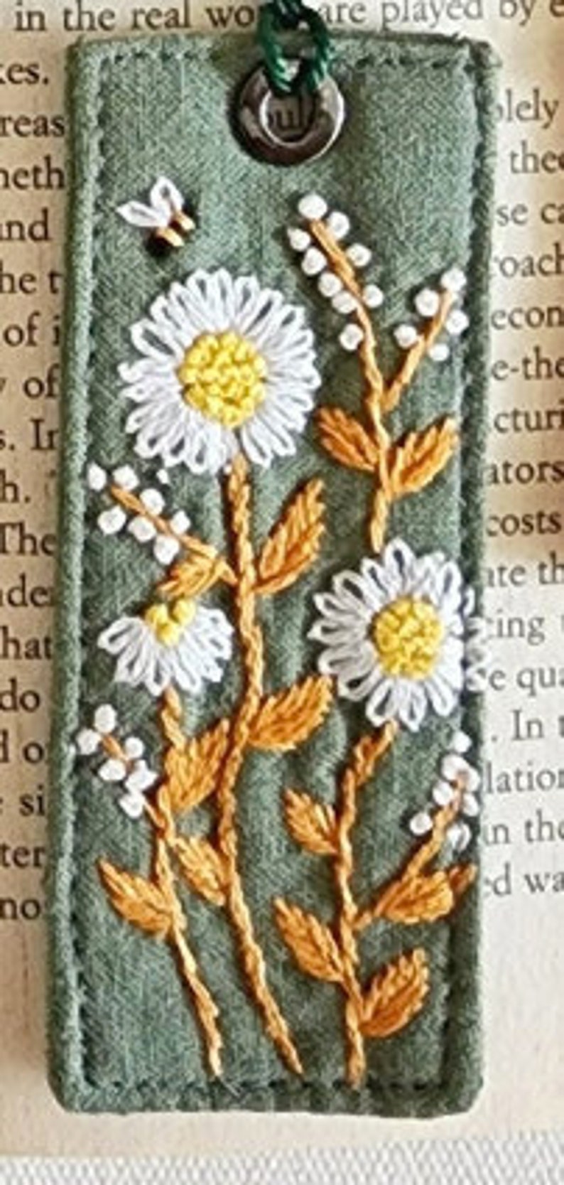 May include: A green fabric bookmark with embroidered white daisies and yellow centers. The stems are embroidered in orange. A small bee is embroidered in the top left corner.