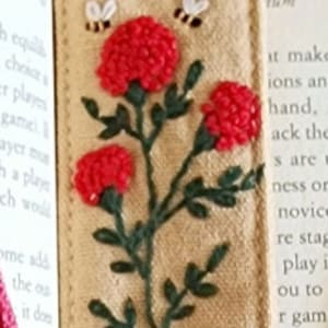 May include: A beige fabric bookmark with a green embroidered stem and red embroidered flowers. Two small bees are embroidered above the flowers. The bookmark has a metal ring at the top.