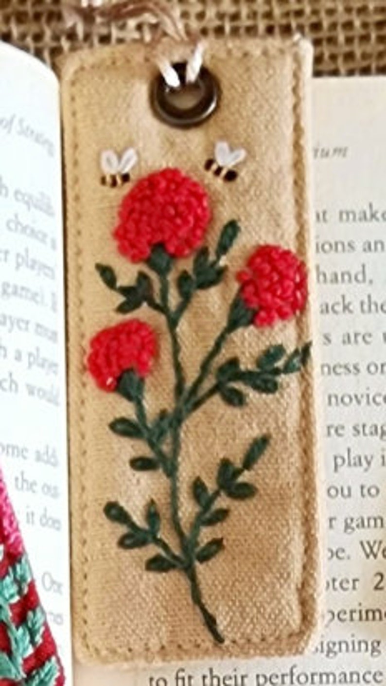 May include: A beige fabric bookmark with a green embroidered stem and red embroidered flowers. Two small bees are embroidered above the flowers. The bookmark has a metal ring at the top.