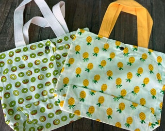 Puede incluir: Dos bolsas de tela reutilizables con asas de tela. Una bolsa tiene un fondo blanco con un patrón de kiwi verde. La otra bolsa tiene un fondo blanco con un patrón de piña amarilla.