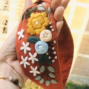 May include: A rust-colored fabric headband with a knotted design, featuring embroidered flowers in shades of yellow, white, blue, and green.
